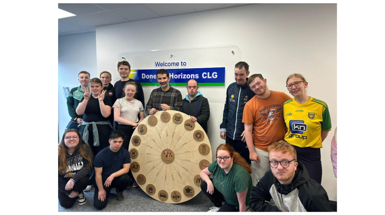 Group of people posing with a large wooden wheel featuring symbols, in front of a sign that says 